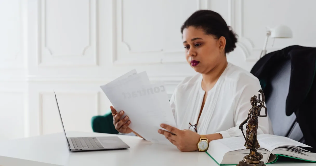 Advogada lendo um contrato em sua mesa com notebook, livros e estátua da Justiça