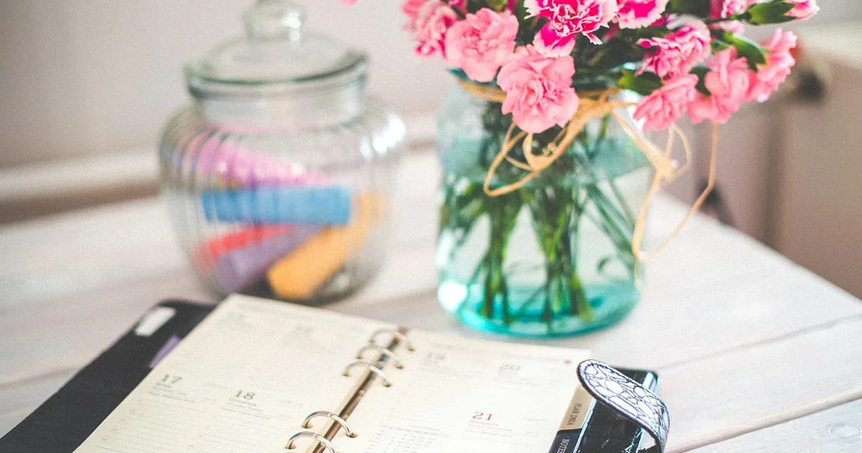 Agenda aberta com dias marcados ao lado de um pote de vidro e um vaso de flores cor-de-rosa